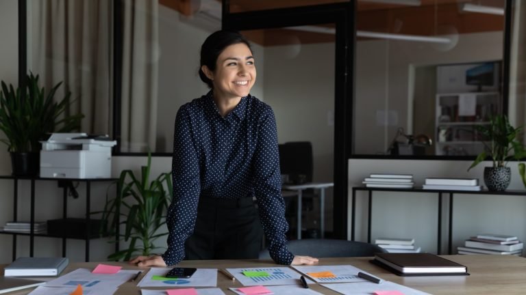 Smiling,Young,Indian,Female,Employee,Stand,At,Desk,In,Office